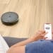 A person uses a smartphone app to control a robotic vacuum on a hardwood floor, showcasing smart home technology.