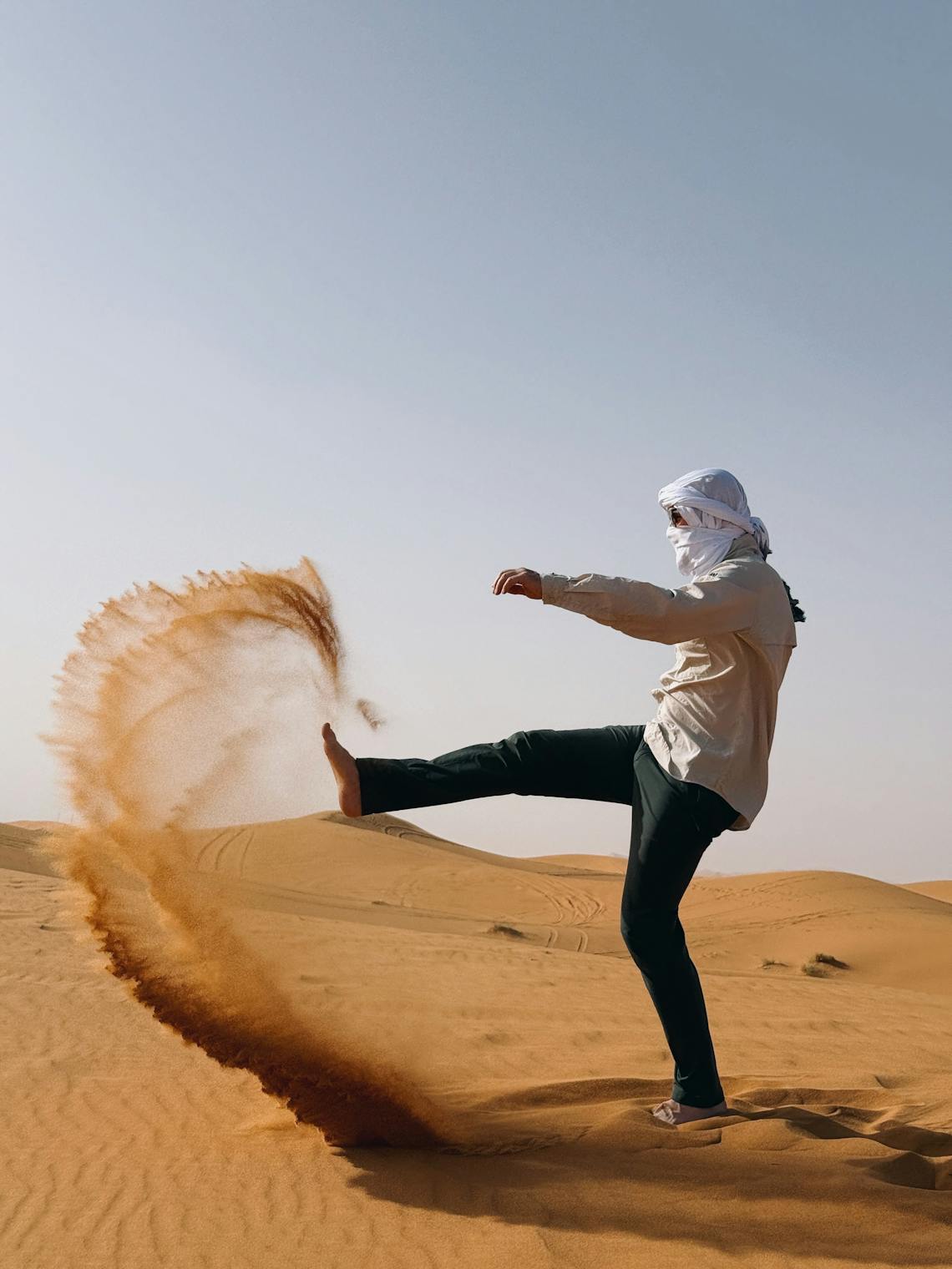 Man in traditional attire kicking sand in a desert, creating a dynamic arc.