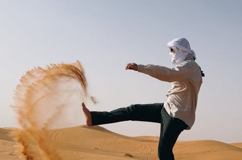 Man in traditional attire kicking sand in a desert, creating a dynamic arc.