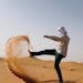 Man in traditional attire kicking sand in a desert, creating a dynamic arc.