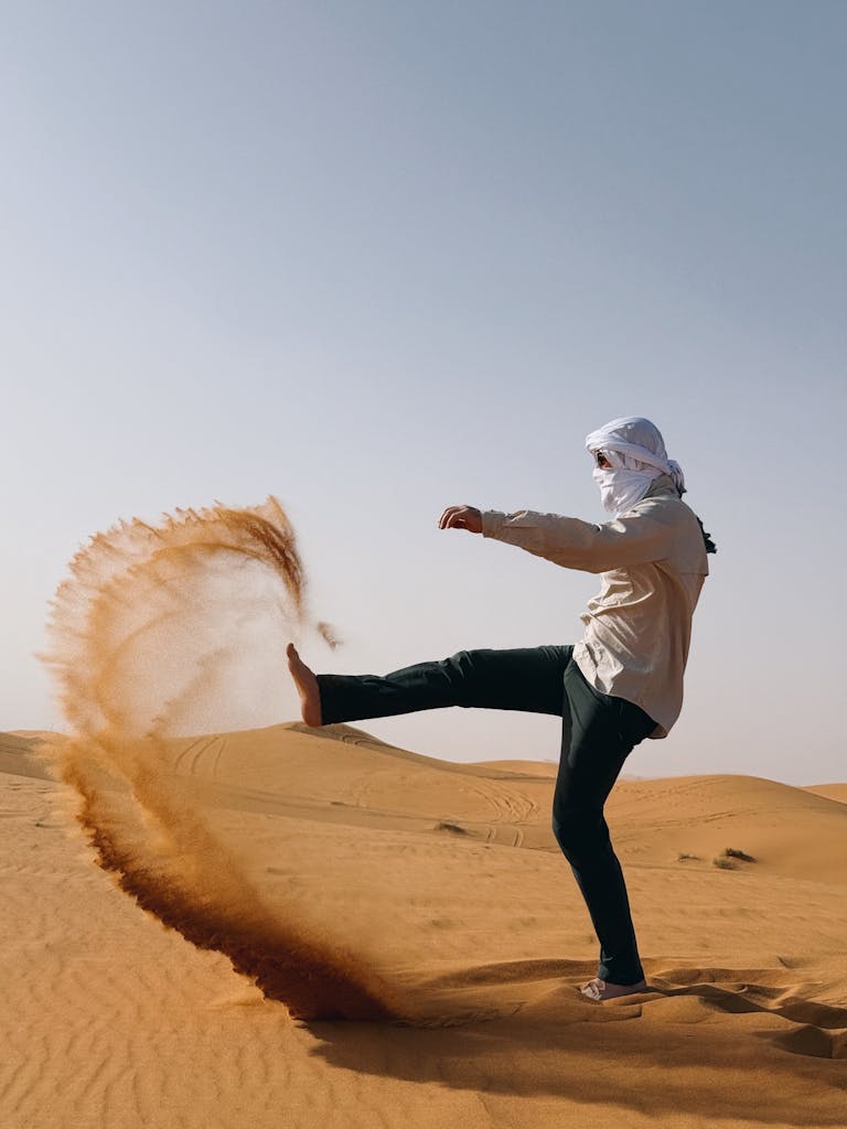 Man in traditional attire kicking sand in a desert, creating a dynamic arc.
